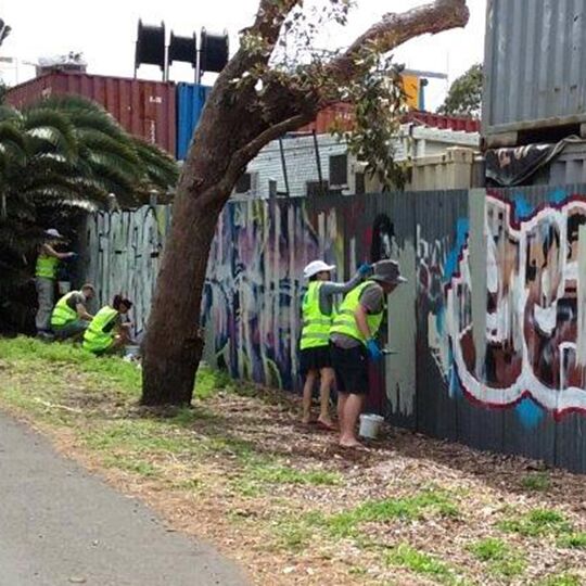 Five council workers in fluorescent yellow vests cleaning graffiti off corrugated iron fences